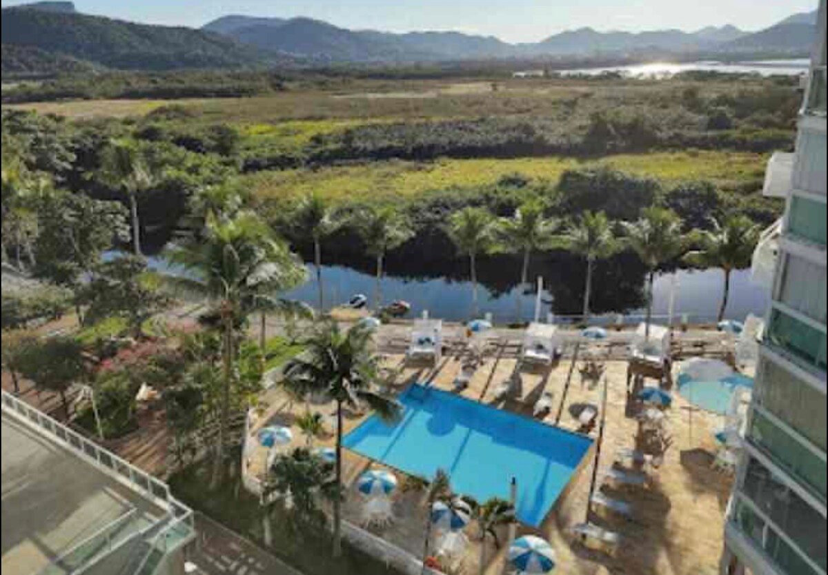 An outdoor pool area is visible, surrounded by sun loungers and umbrellas. Lush greenery and palm trees frame the space, leading down to a tranquil body of water. Scenic mountains are seen in the distance, with clear skies and sunlight enhancing the view.