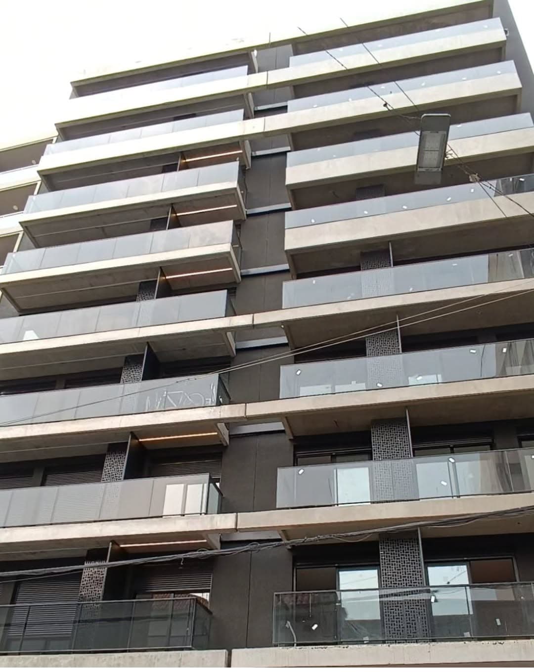 A modern multi-story building features a sleek design with a combination of dark concrete and glass balconies. The structure showcases multiple levels, each equipped with railings, allowing for views of the surrounding area.