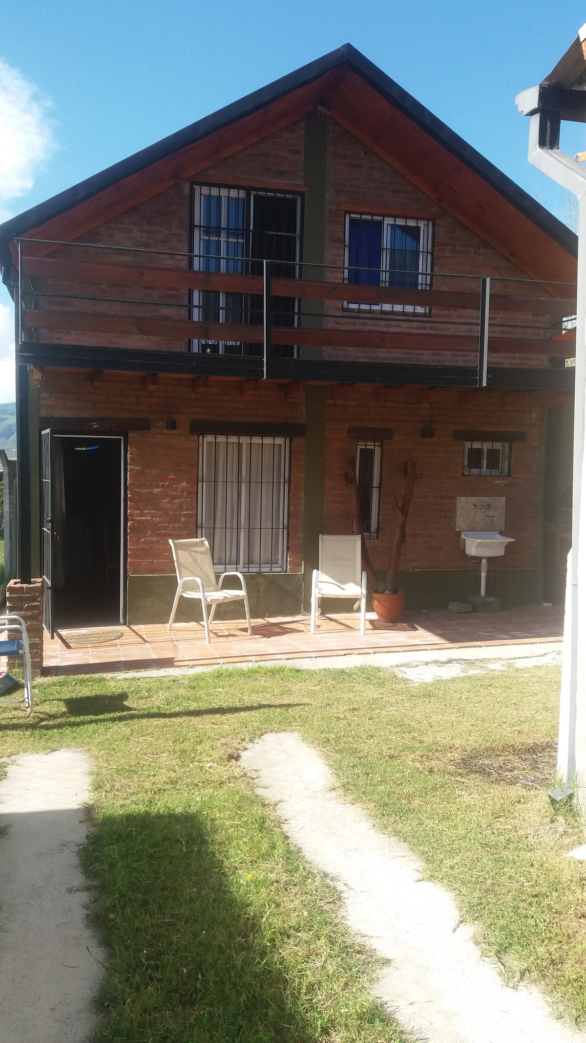 A two-story house features a blend of brick and wood accents under a clear blue sky. A small balcony is visible on the upper level, and two white chairs are positioned on the patio area. A grassy path leads from the foreground to the entrance.