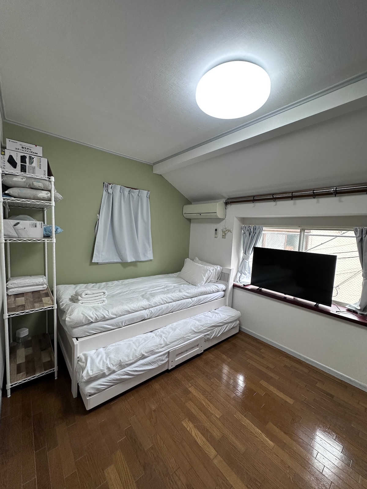 A cozy sleeping area features a twin bed with white linens placed against a green accent wall. A large window allows natural light to enter, complemented by light curtains. A TV is positioned near the window, and a shelf holds neatly stacked towels.