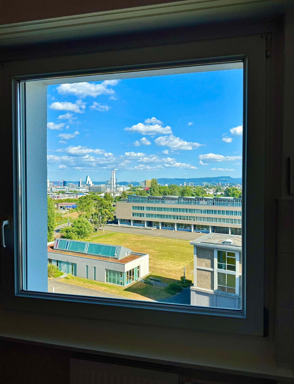 A view through a window captures a vibrant outdoor scene featuring a clear blue sky dotted with fluffy clouds. The foreground includes green spaces and modern buildings, while distant mountains create a scenic backdrop.