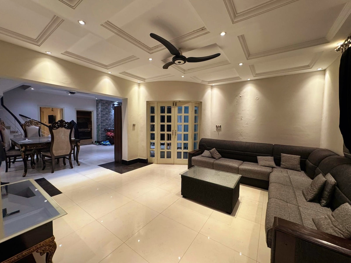 A spacious lounge area features a large L-shaped grey sofa and a modern coffee table, surrounded by soft lighting that emphasizes the coffered ceiling. Double glass doors lead to another room, while a dining area with elegant chairs is visible in the background.