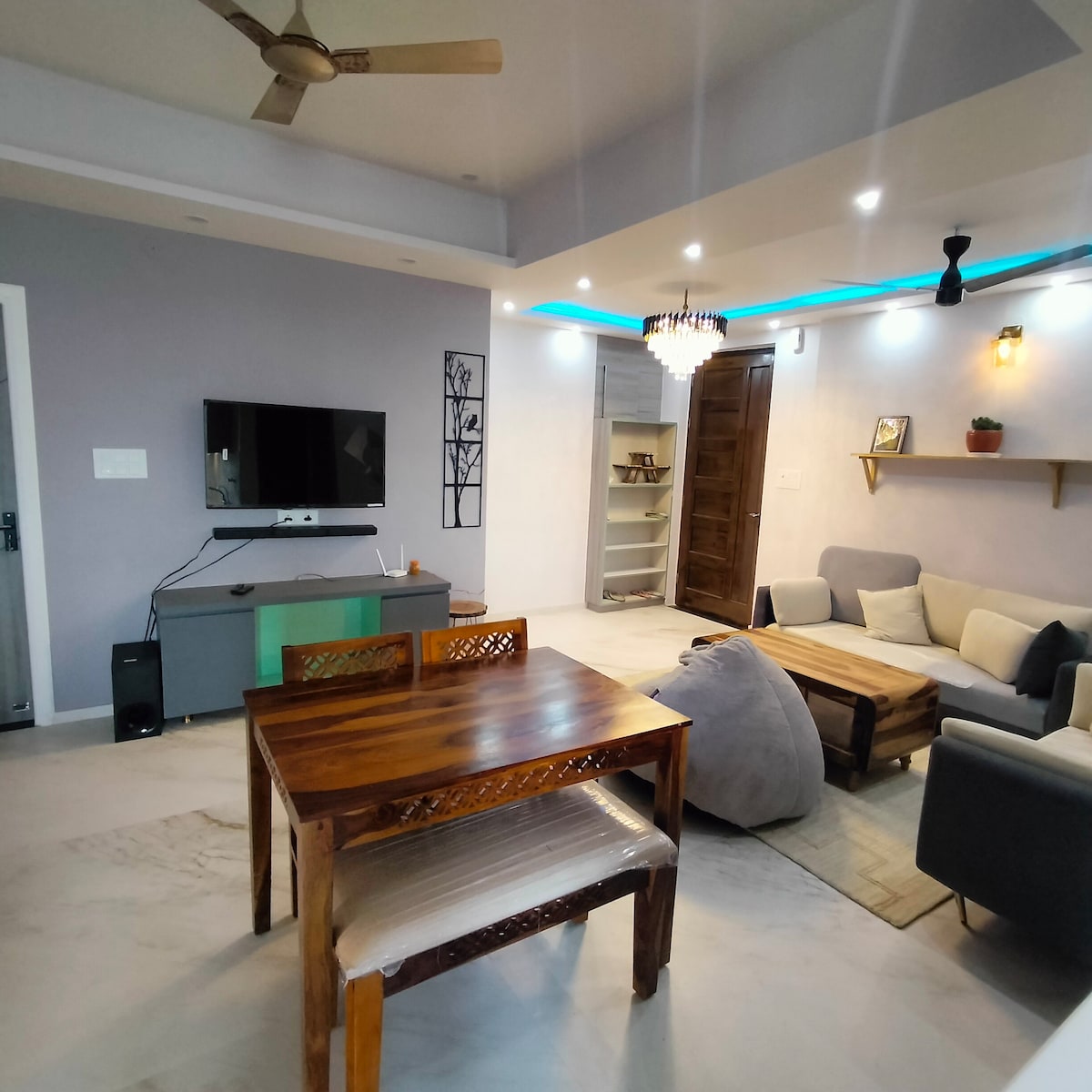 A welcoming living space is presented, featuring a wooden dining table and comfortable seating arrangements. A modern television is mounted on the wall opposite a sleek coffee table. Soft lighting is provided by ceiling fixtures, enhancing the relaxed atmosphere of the room.