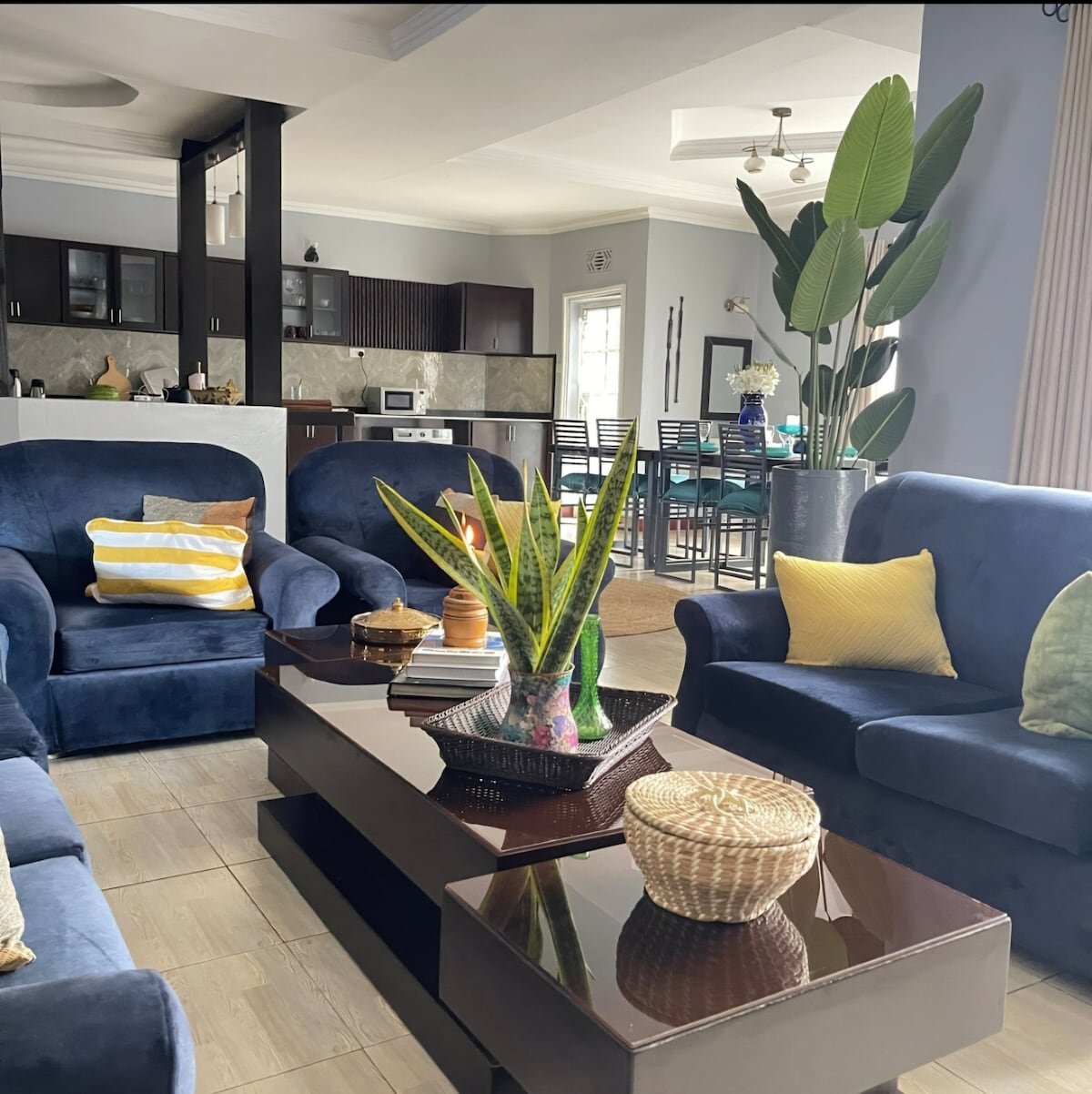 A spacious living area features two comfortable blue sofas and a dark coffee table adorned with a patterned plant arrangement. Natural light filters through large windows, illuminating the room. In the background, a dining space is visible with a modern table and chairs.