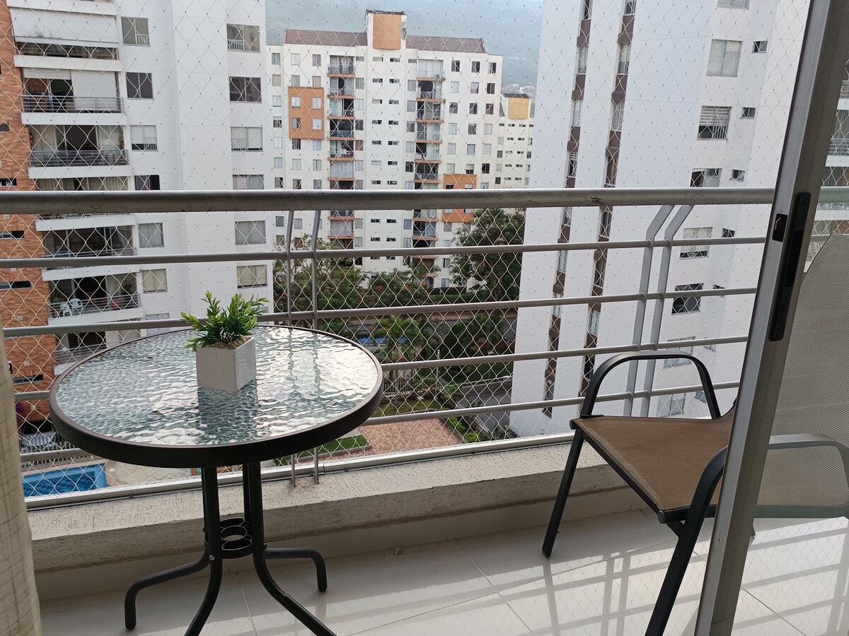 A private balcony is equipped with a small round table and a single chair, overlooking a residential area. The view includes surrounding buildings and greenery, with a hint of a swimming pool in the distance. Decorative plants are placed on the table.