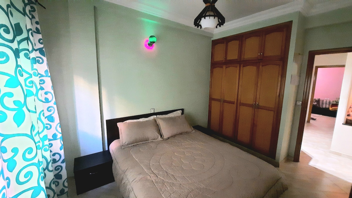 A spacious bedroom is presented with a neatly made bed featuring a light-colored quilt. A wardrobe with wooden doors is positioned against the wall. An accent lamp provides colorful lighting, complementing the room's soft green walls and patterned curtains that frame a window.