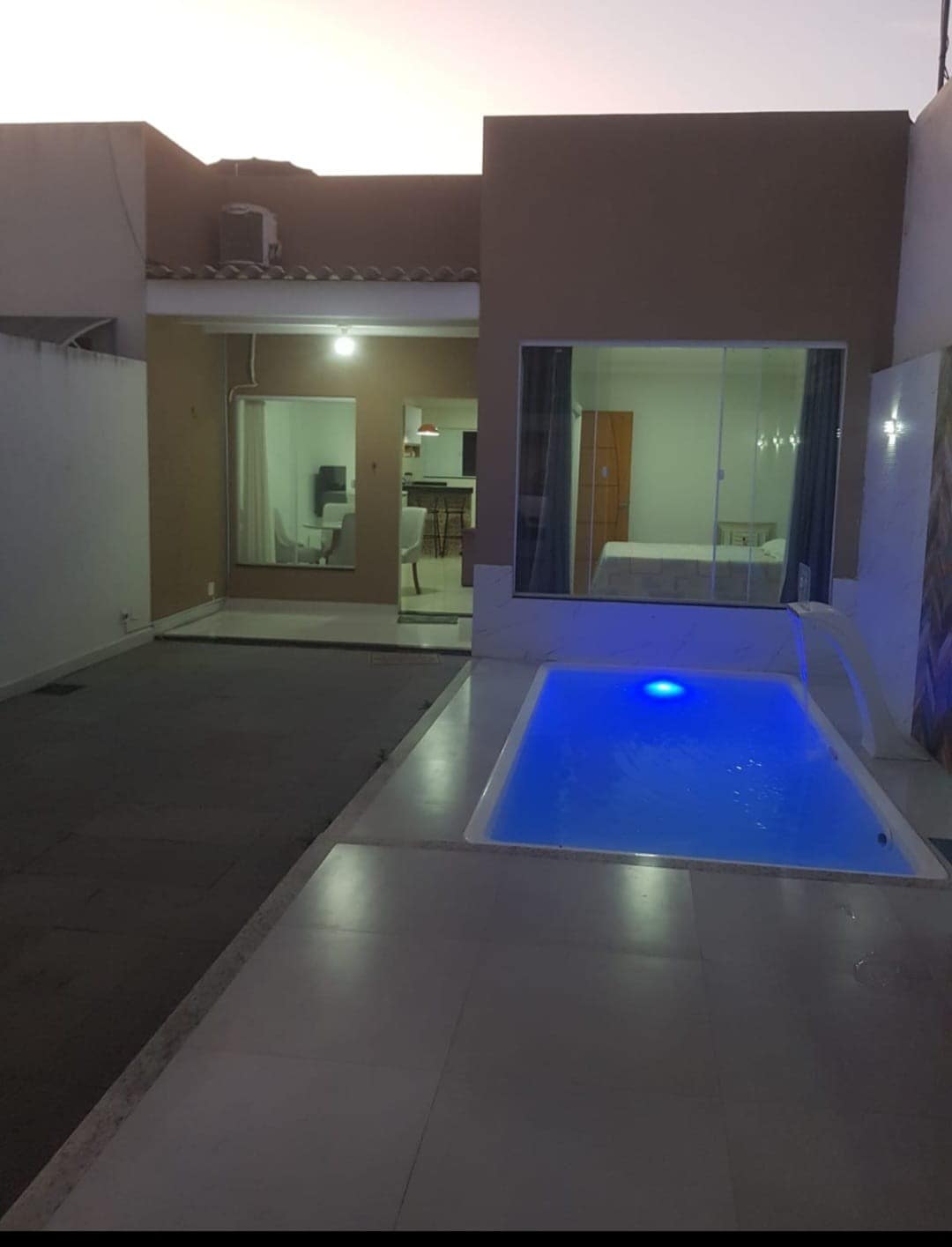 A modern outdoor area features a small pool with blue lighting. One side provides a view into the living space, while the other showcases a dining area through large glass doors. The space is minimally furnished and well-lit, with a neutral color palette.