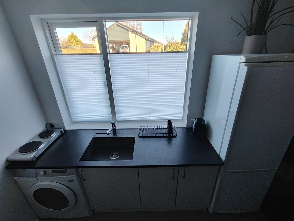 A modern kitchen is equipped with essential appliances, including a washer and a refrigerator. A dark countertop offers space for meal preparation, while a sink is positioned near a window. Natural light enters through the covered glass, providing a bright and airy feel.