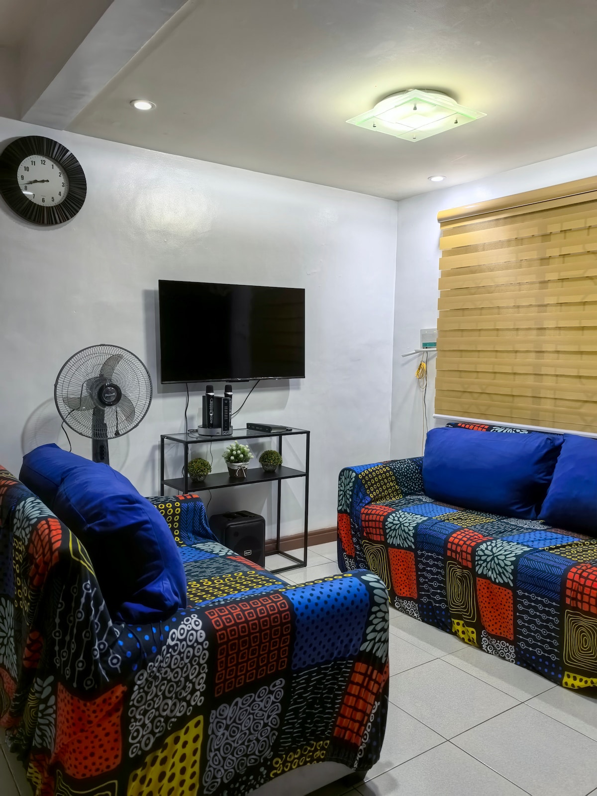 A cozy living area features two patterned sofas with blue cushions, arranged around a black coffee table. A wall-mounted television is visible, accompanied by a small sound system. Natural light filters through bamboo blinds, creating a relaxed atmosphere.