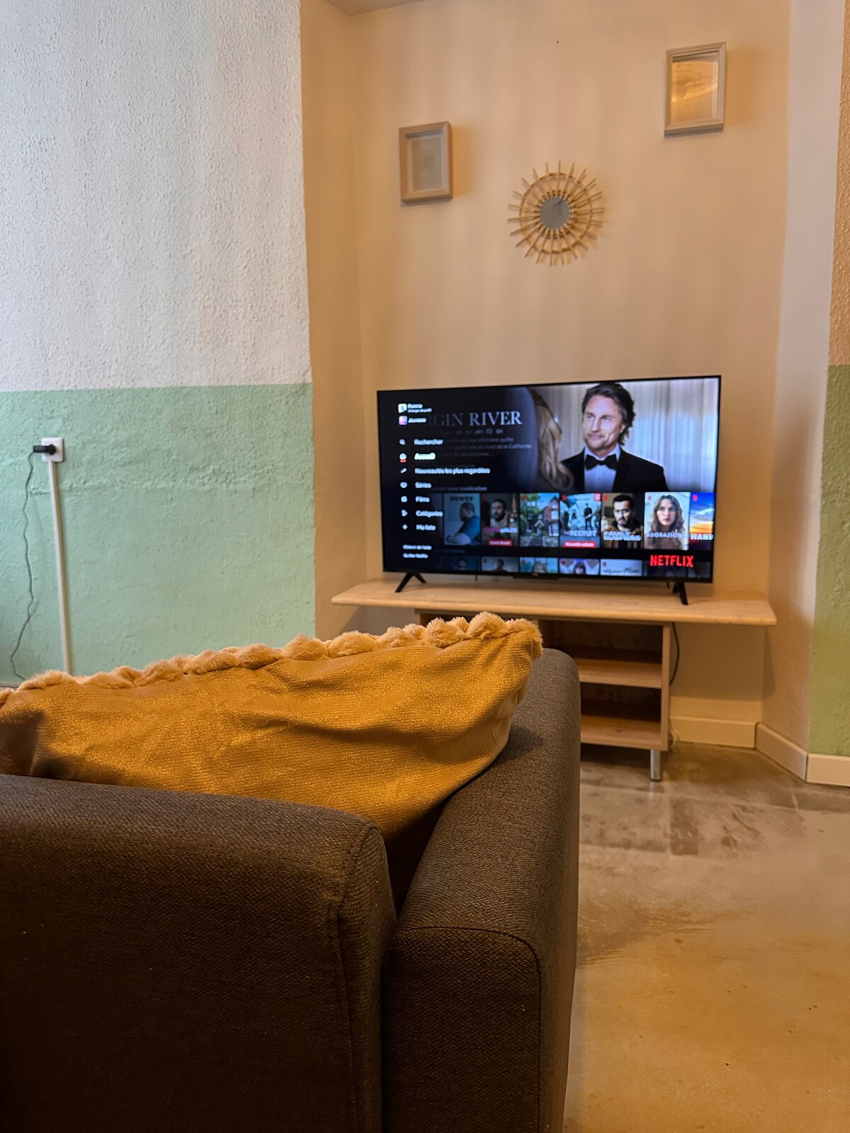 A comfortable living area features a gray sofa adorned with a soft, folded blanket. A wall-mounted television is positioned on a light wooden stand, displaying a streaming interface. The wall behind the TV is painted in a two-tone color scheme, adding a stylish touch.