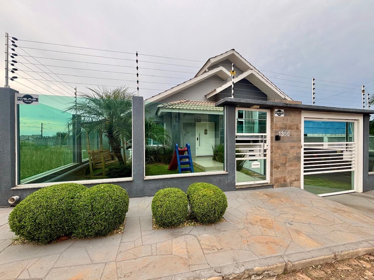 The exterior of the house features large glass windows that provide visibility into a welcoming entryway. A manicured garden with rounded hedges lines the walkway, while a colorful children's play structure and seating area can be seen through the transparent façade.
