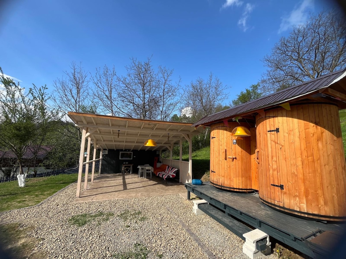 A covered outdoor area features comfortable seating with striped cushions and hanging lanterns. Two wooden barrel structures are positioned nearby, offering rustic charm. Green grass and bare trees enhance the natural setting under a clear blue sky.