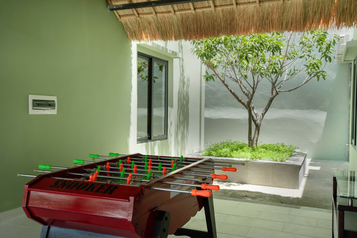 A snooker table in a brightly lit area features colorful cues, set against green walls. Natural light filters through nearby windows, illuminating a small tree and greenery in the background, enhancing the inviting space for leisure activities.