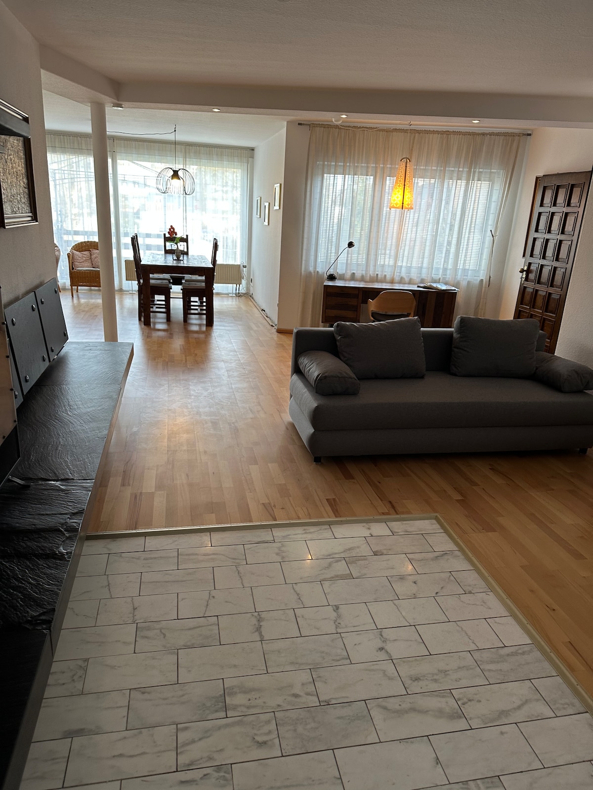 A spacious living area is shown, featuring a comfortable gray sofa and a modern tile floor. Natural light filters through large windows with sheer curtains. A dining table and chairs are visible in the background, along with a wooden sideboard, enhancing the open layout.