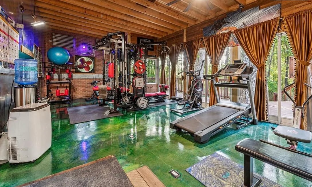 A fitness room is equipped with various exercise machines, including a treadmill and weight bench. Bright windows provide natural light, and wooden beams enhance the ceiling. Vibrant green flooring adds a unique touch, while curtains offer privacy when needed.