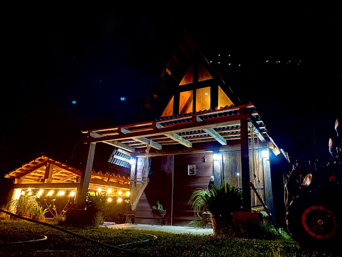 The exterior of a charming cabin is illuminated at night, showcasing its wooden architecture and welcoming facade. Soft lights accentuate details of the structure and surrounding greenery, creating a peaceful atmosphere. The sloped roof and overhang provide a cozy appeal against the night sky.