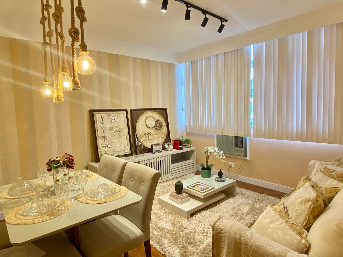 A dining area features a glass table set for four, complemented by muted gray chairs. Warm light is provided by hanging bulb fixtures. A beige couch is positioned nearby, creating a cozy atmosphere, while large windows allow natural light to fill the space.