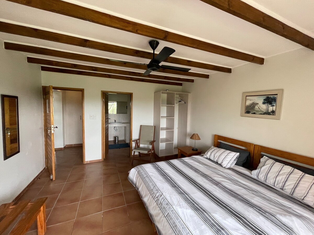 A spacious bedroom is featured with a wooden beam ceiling, two structured beds adorned with striped linens. A small lamp sits on a bedside table, and light enters through a nearby window. A closet and entrance to a bathroom complete the room's layout.