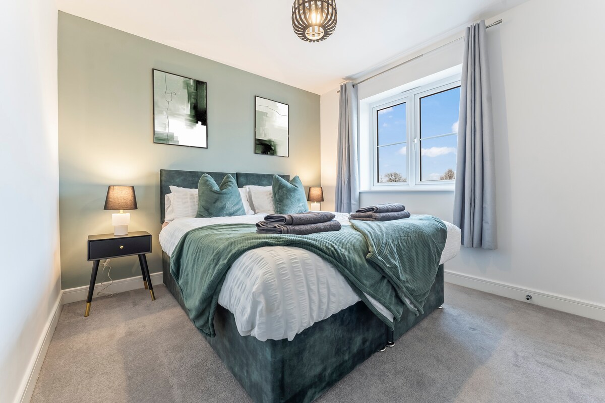 A well-lit bedroom features a spacious bed dressed in soft linens and complemented by decorative pillows. Two nightstands, each with a lamp, are situated beside the bed. A large window allows natural light to illuminate the room, while light gray curtains frame the view.