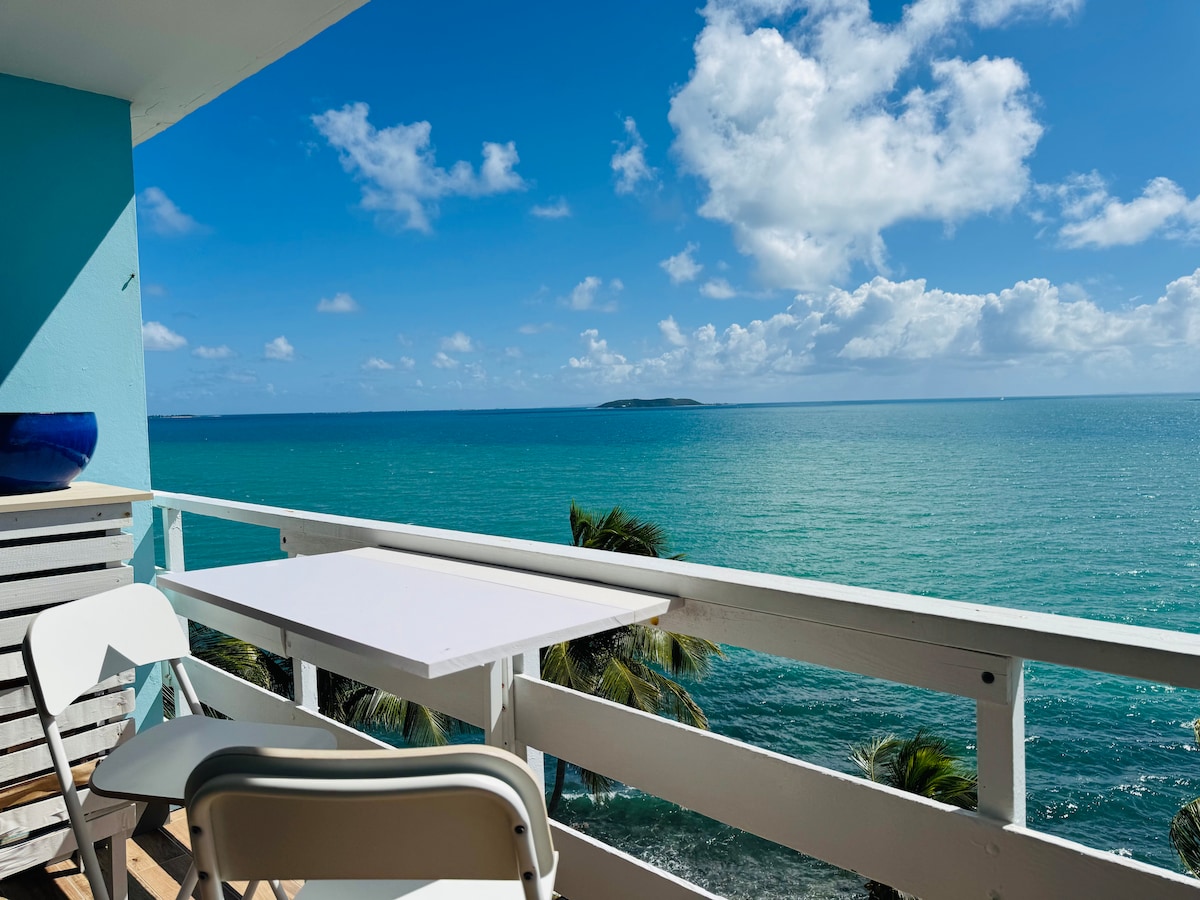 A balcony view offers expansive outlooks of the turquoise sea under a partly cloudy sky. A small white table and two chairs are positioned along the railing, creating a space to enjoy the serene surroundings and the gentle movement of palm trees in the breeze.