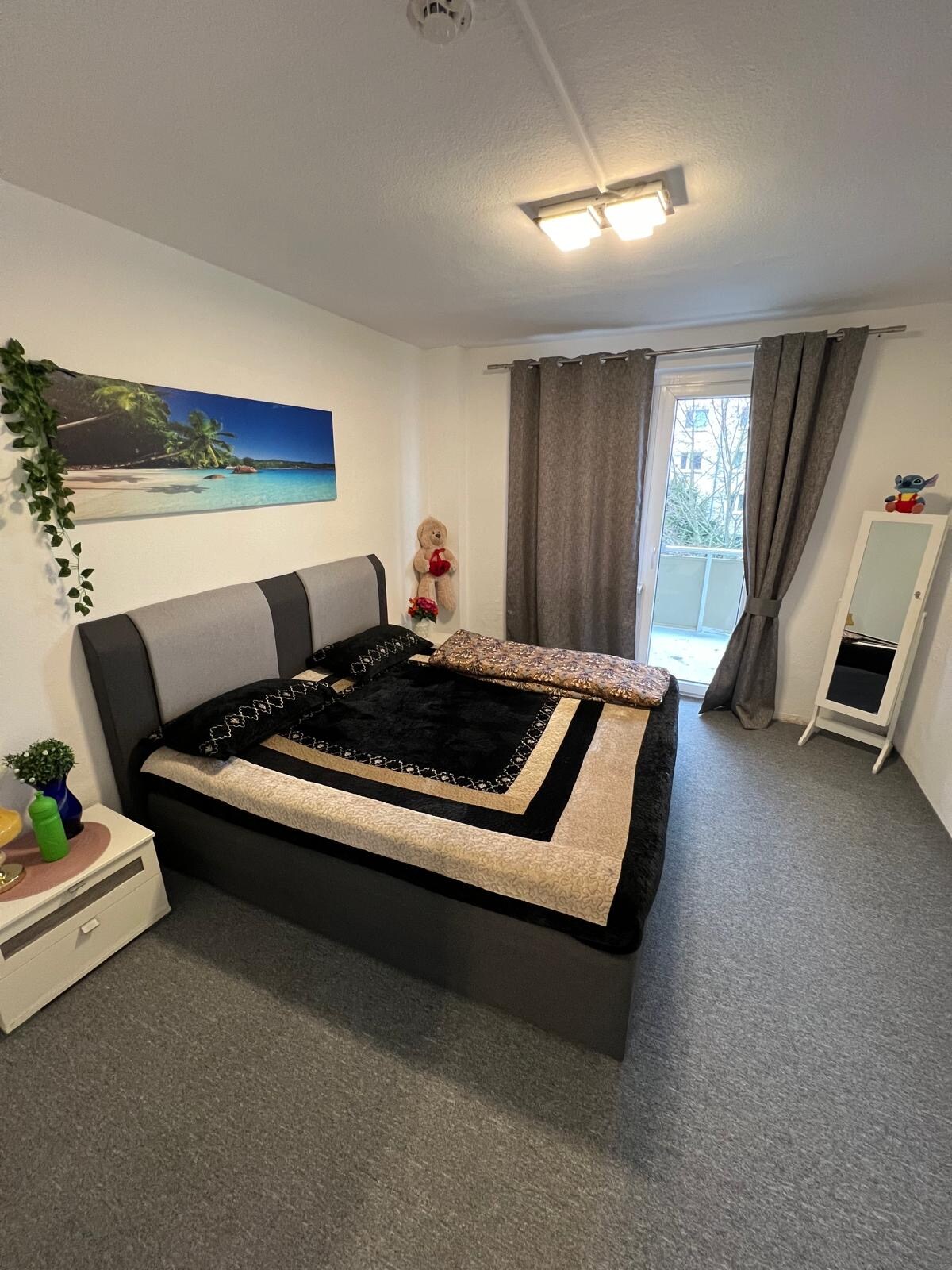 A cozy bedroom features a spacious bed with dark bedding and textured throws. A piece of art depicting a beach hangs above the bed. Natural light enters through a nearby balcony door, complemented by soft gray curtains. A white wardrobe and small nightstand add functionality to the space.