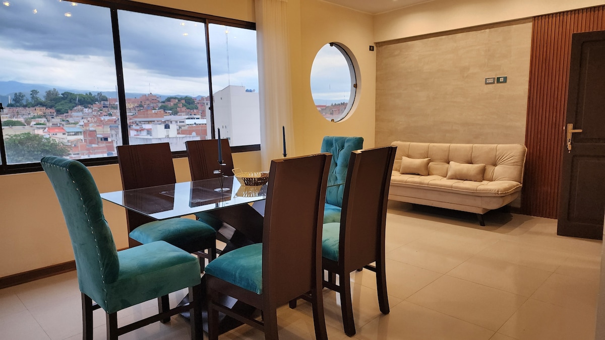 A spacious dining area is furnished with a glass table surrounded by six upholstered chairs. A comfortable sofa is positioned against the wall, while large windows offer a view of the surrounding cityscape and natural light fills the space.