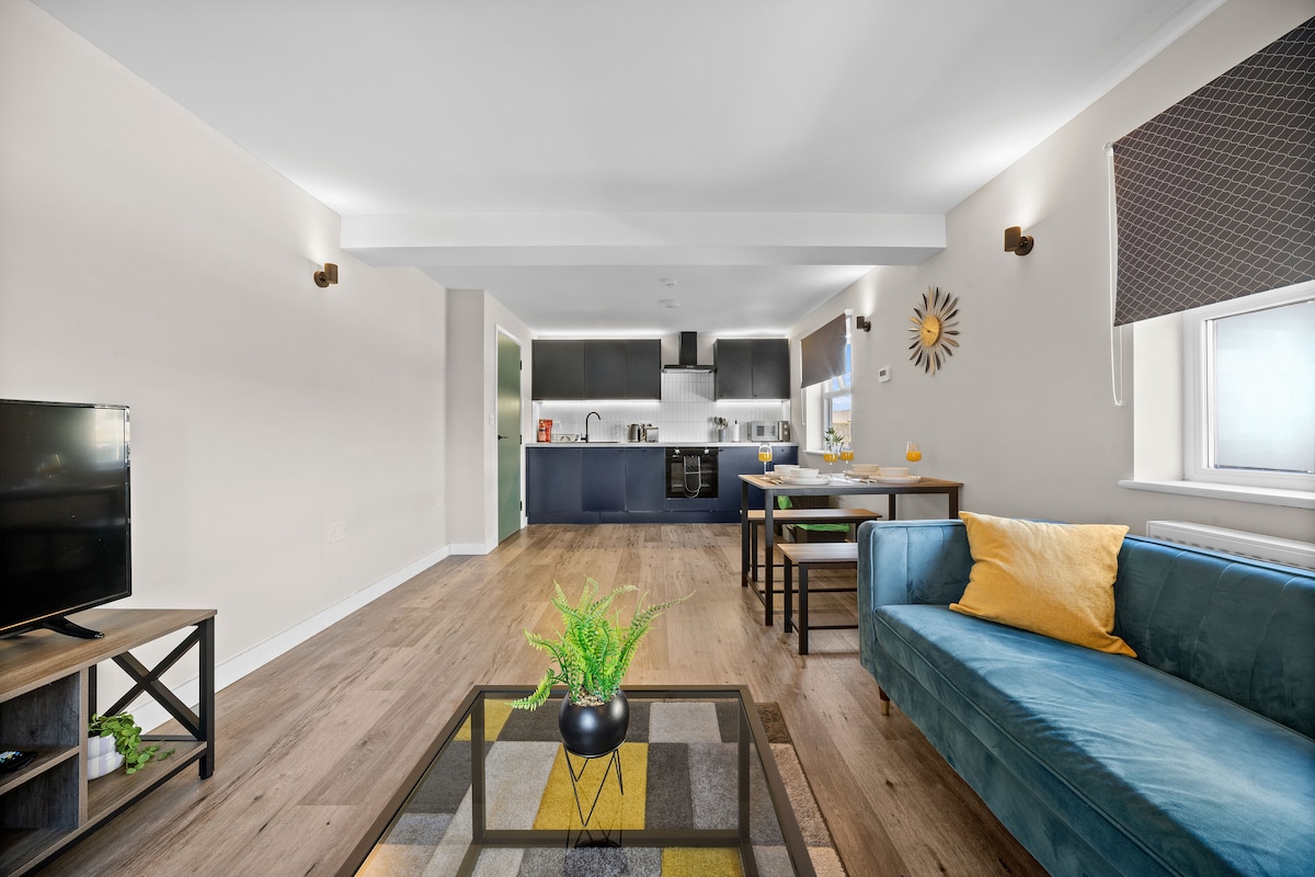 A well-lit living area features a comfortable blue sofa with a yellow cushion. A coffee table with a patterned surface sits at the center, surrounded by warm wooden flooring. A dining table for four is visible, along with a modern kitchen in the background.