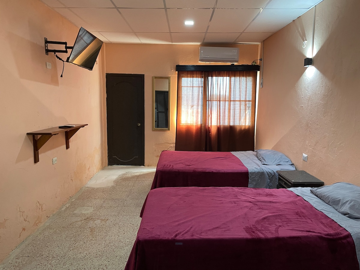 The room features two spacious beds dressed in deep red linens, positioned side by side. A wall-mounted TV is visible, along with a small nightstand beside each bed. Natural light is filtering through partially open brown curtains, illuminating the room's neutral-toned walls.