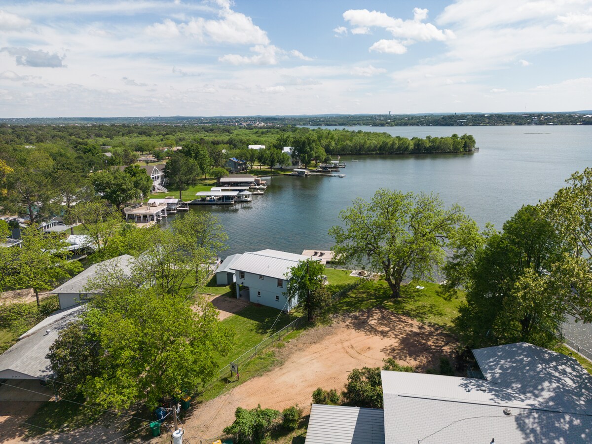 Aerial view of the tranquil Lake LBJ coastline is displayed, showcasing lush green trees and nearby homes. The clear water is visible, along with boats docked at piers. The landscape encompasses both natural and residential elements, contributing to the serene waterfront setting.