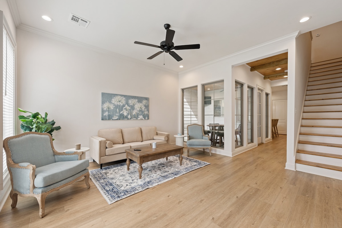 A light-filled living area features a comfortable beige sofa, two accent chairs, and a wooden coffee table. A large area rug adds warmth to the space. Large windows allow natural light, and a ceiling fan enhances airflow. Staircase and dining area are visible in the background.