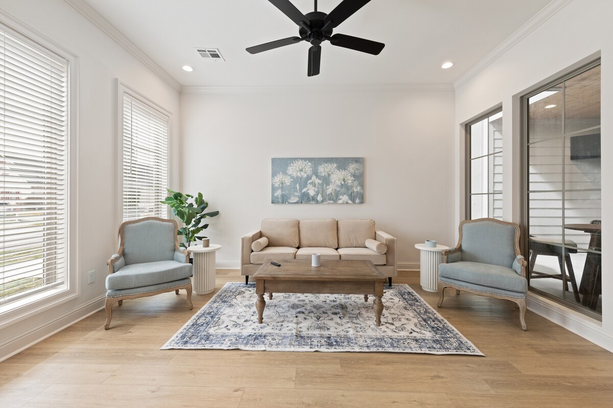 A comfortable living area features a light-toned sofa and two upholstered chairs, creating a welcoming atmosphere. Natural light floods in through large windows, complemented by a decorative rug and a coffee table placed in the center. Indoor plants add a touch of greenery to the space.