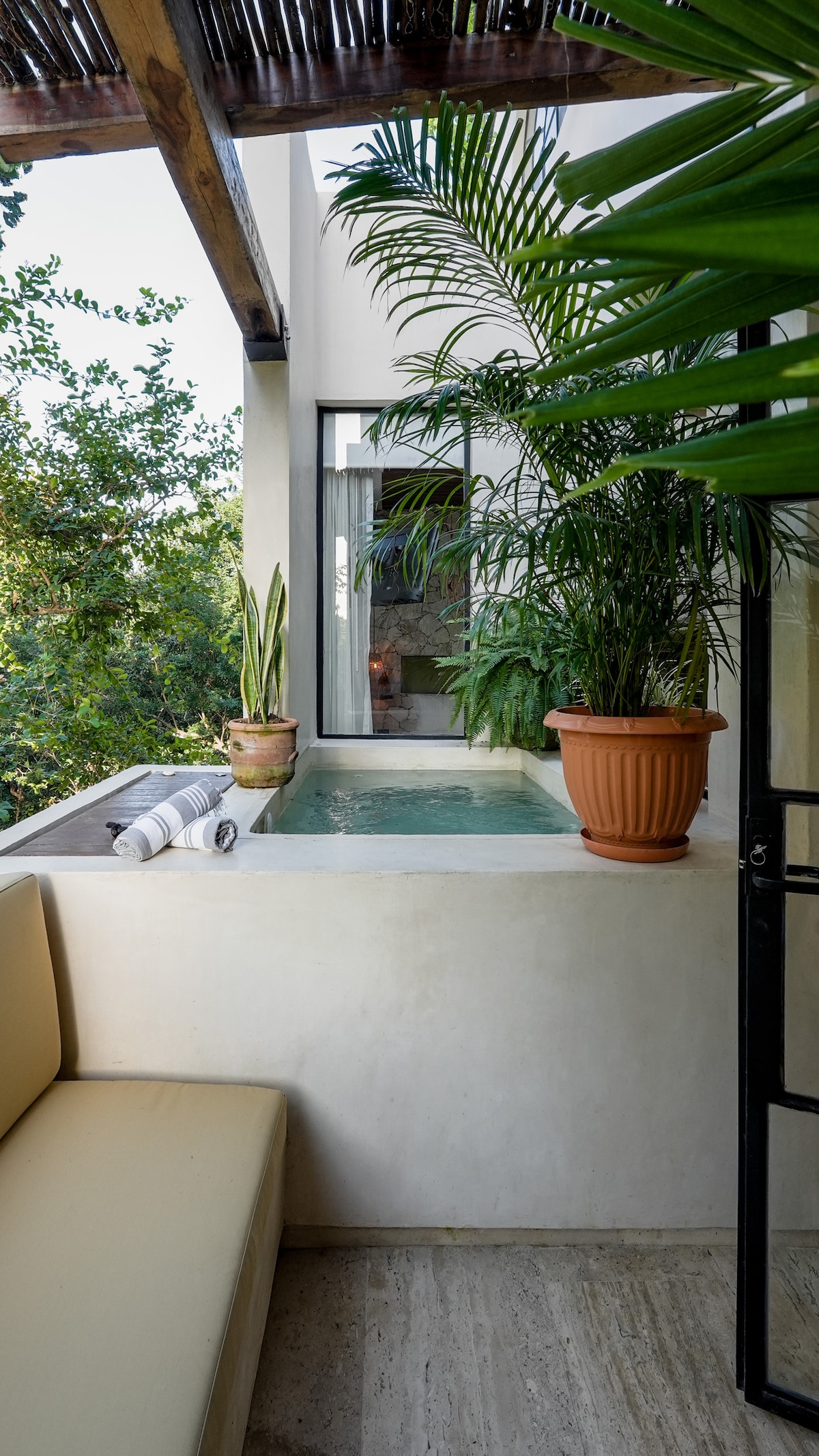 A private plunge pool is situated beside a seating area, surrounded by lush greenery. Potted plants add a natural touch, creating a serene atmosphere. A window provides a view of the outdoor space, and rolled towels are neatly placed nearby, enhancing the inviting setting.