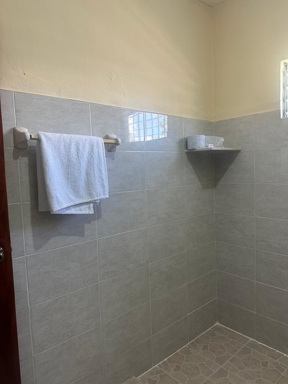 A clean and functional shower area is presented, featuring grey tiled walls and a light-coloured floor. A towel hangs on a wall-mounted rack, while a small corner shelf holds toiletries. Natural light enters from a window positioned high on the wall.