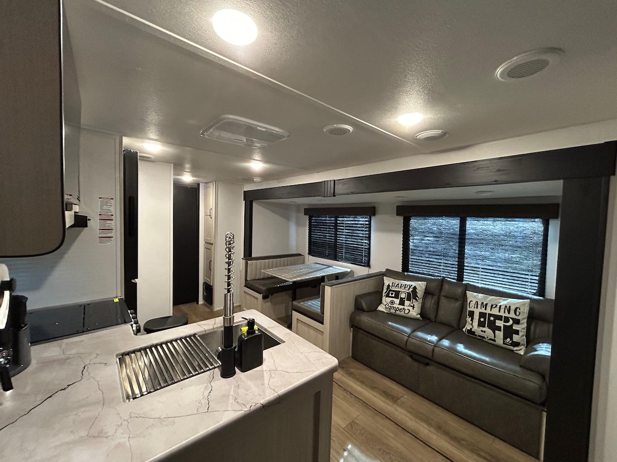 The interior of the camper showcases a sleek kitchen area with a marble countertop and modern appliances. A cozy living space is seen with a dark sofa adorned with decorative cushions, complemented by a dining area with a built-in bench. Large windows provide natural light.