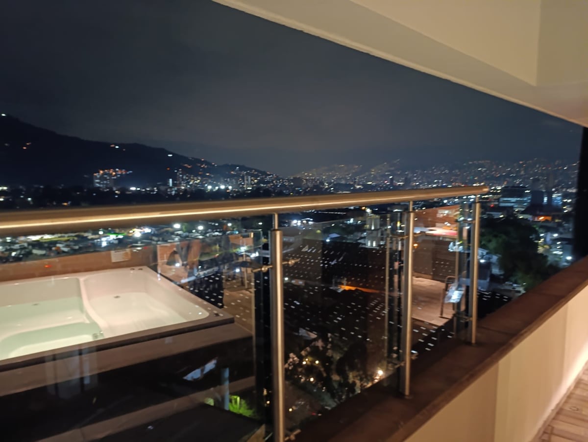 A balcony view showcases a city skyline illuminated at night, with twinkling lights across the landscape. A spa is visible on the left, and a sleek glass railing enhances the view of the scenery beyond.