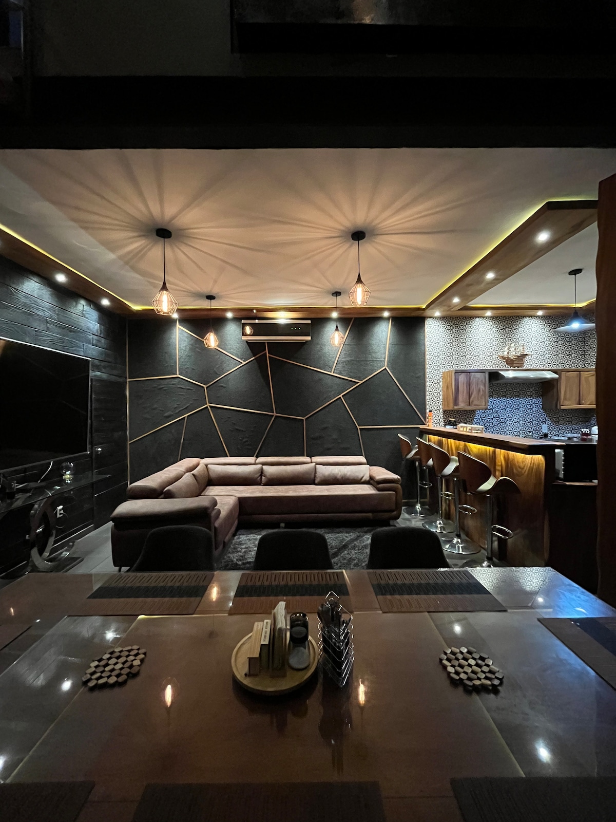 A modern living area features a spacious sectional sofa arranged on a textured rug. A sleek dining table is visible in the foreground, accompanied by black chairs. Atmospheric lighting highlights the geometric wall design, creating a stylish and inviting ambiance.