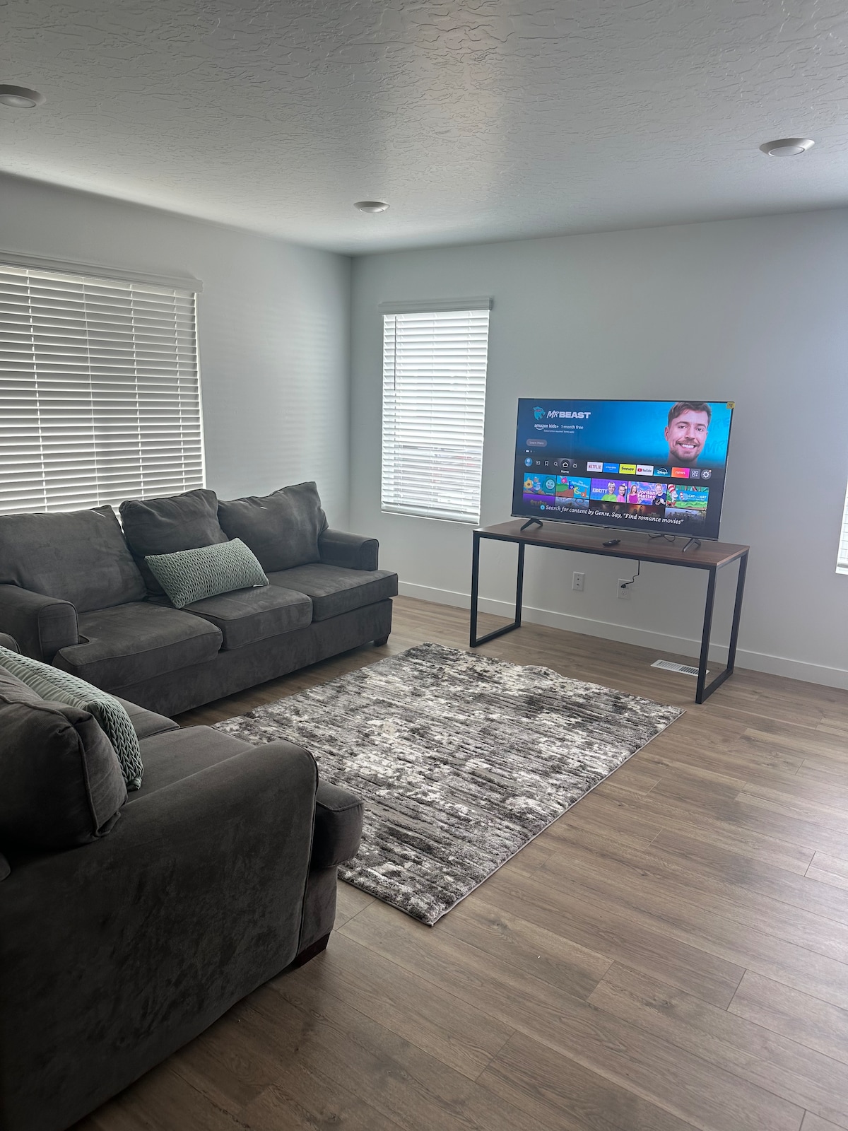 A bright living area features two dark grey sofas positioned near large windows, allowing ample natural light. A flat-screen television is mounted on the wall beside a sleek console table. A textured area rug adds warmth to the wooden flooring.