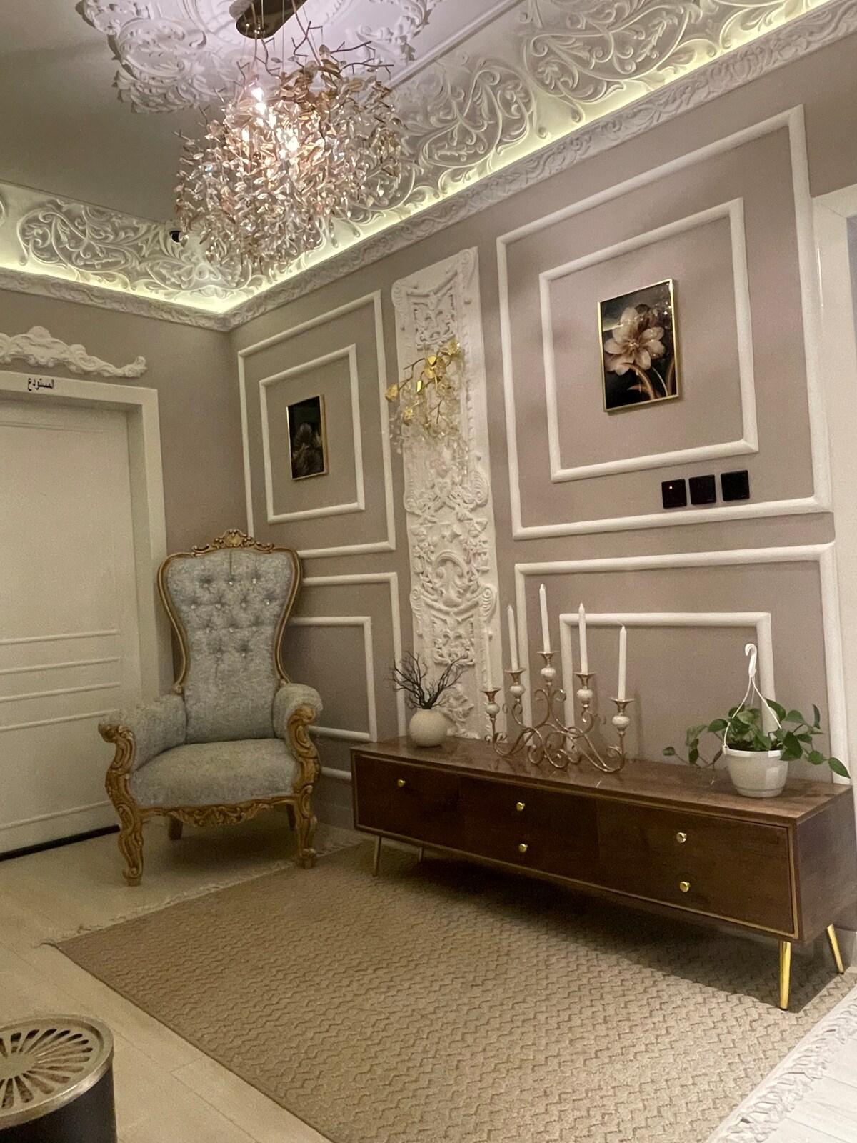 A stylish entryway is showcased, featuring an elegant high-backed chair and a sleek wooden console table. The walls are adorned with intricate moldings and framed art. Soft lighting is provided by a decorative ceiling fixture, enhancing the warm tone of the space.