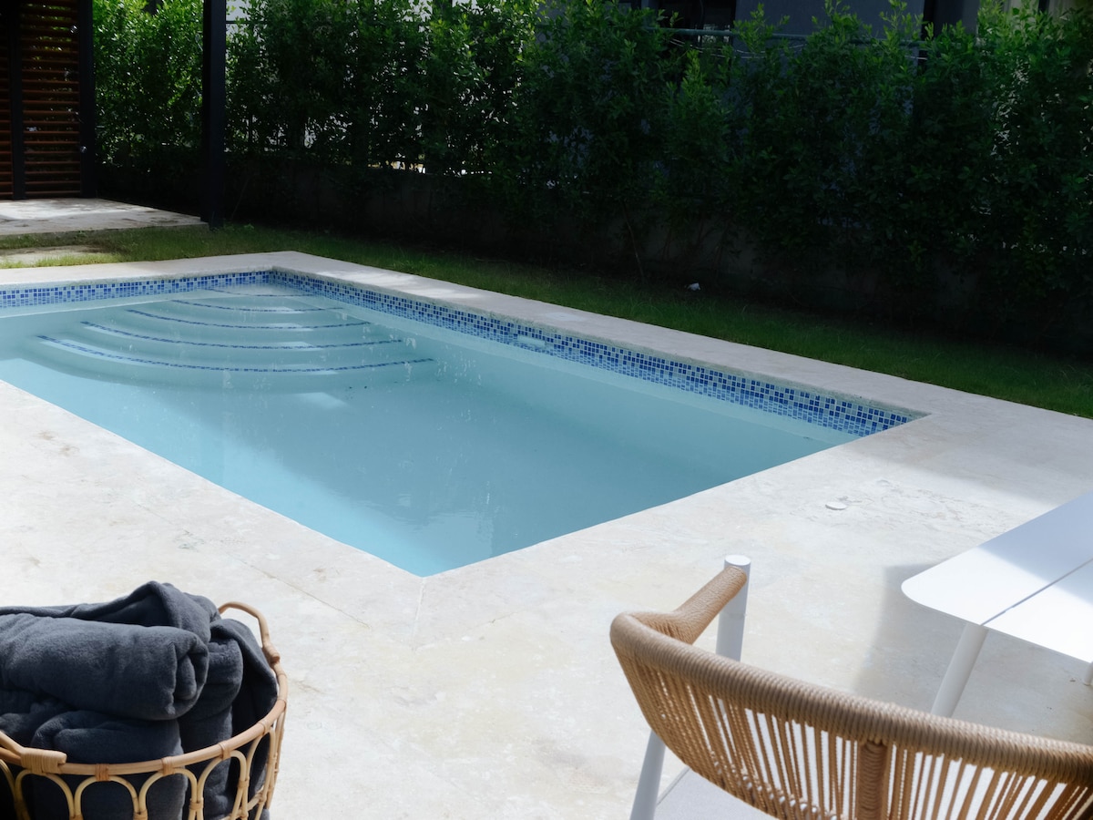A private pool is surrounded by a spacious patio. A few towels are neatly stacked in a woven basket, while a modern chair offers a spot for relaxation. Lush greenery forms a natural border around the pool area, enhancing privacy.