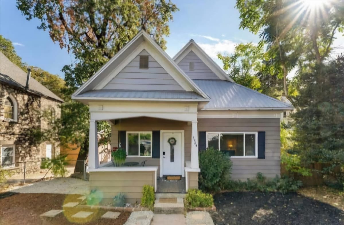 The front exterior of the house showcases a charming design with a light gray facade and a welcoming porch. Green landscaping and a path lead to the entrance, while trees provide a natural backdrop. Sunlight filters through the leaves, illuminating the welcoming atmosphere.