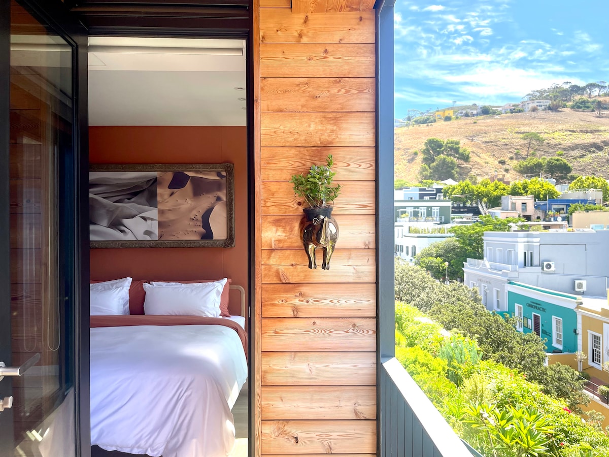A balcony view reveals a spacious bedroom with a plush bed framed by warm wooden paneling. On the wall, a planter adds greenery, while a scenic backdrop of hills and colorful buildings is visible through the wide glass doors.