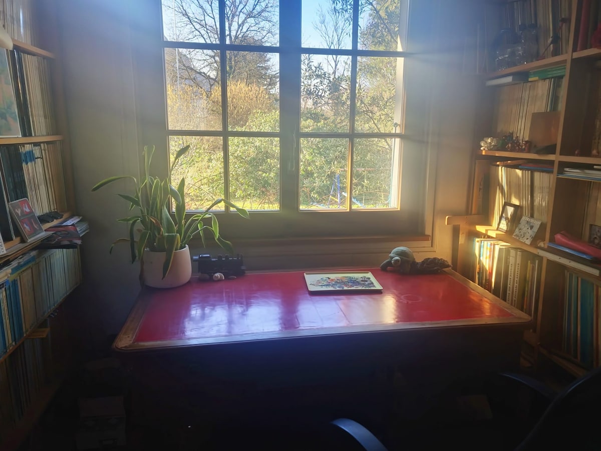 A well-lit workspace features a wooden desk topped with a flat surface and a potted plant. Shelves lined with books and collectibles surround the area. Natural light streams through a large window, providing a view of greenery outside.