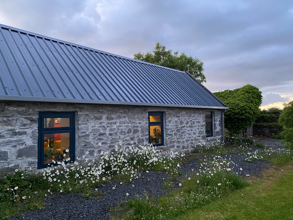 The exterior of the stone cottage is framed by a vibrant garden of wildflowers, with a gravel path leading to the entrance. Large windows reflect warm interior lights, hinting at the inviting atmosphere within. The roof is clad in metal, providing a modern touch to the rustic structure.