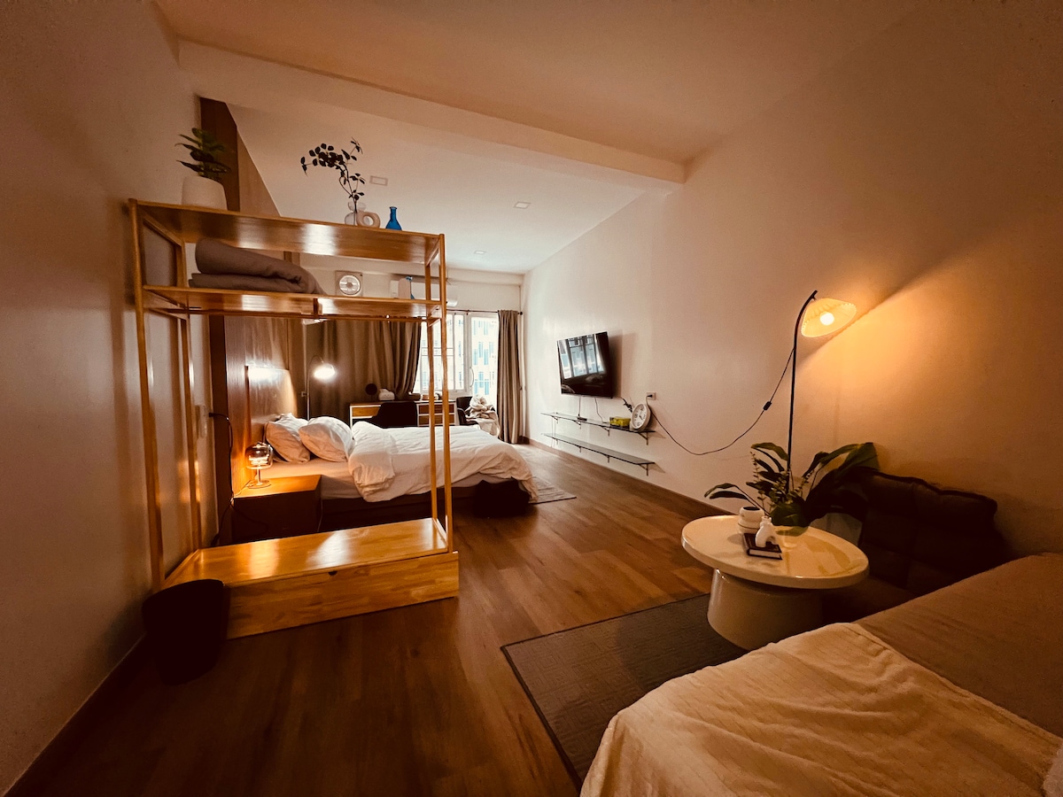 A modern studio features a wooden double bed with soft linens and a cozy seating area with cushions. A small round table is positioned next to a plant. Natural light filters through a window, illuminating the space, which includes a television mounted on the wall.