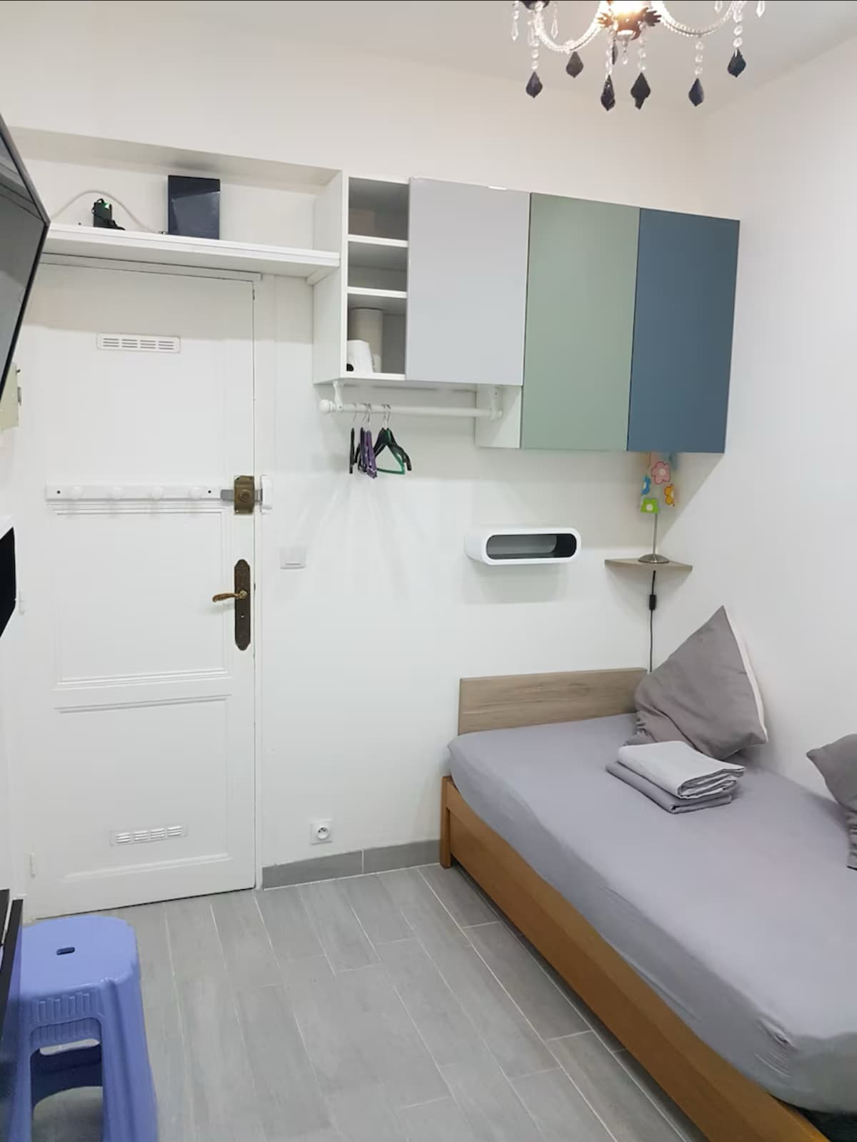 A compact living space features a spacious bed with neatly arranged bedding and a small folding stool nearby. A TV and storage shelves are mounted on the wall. Light colors are used throughout, enhancing the room's brightness.