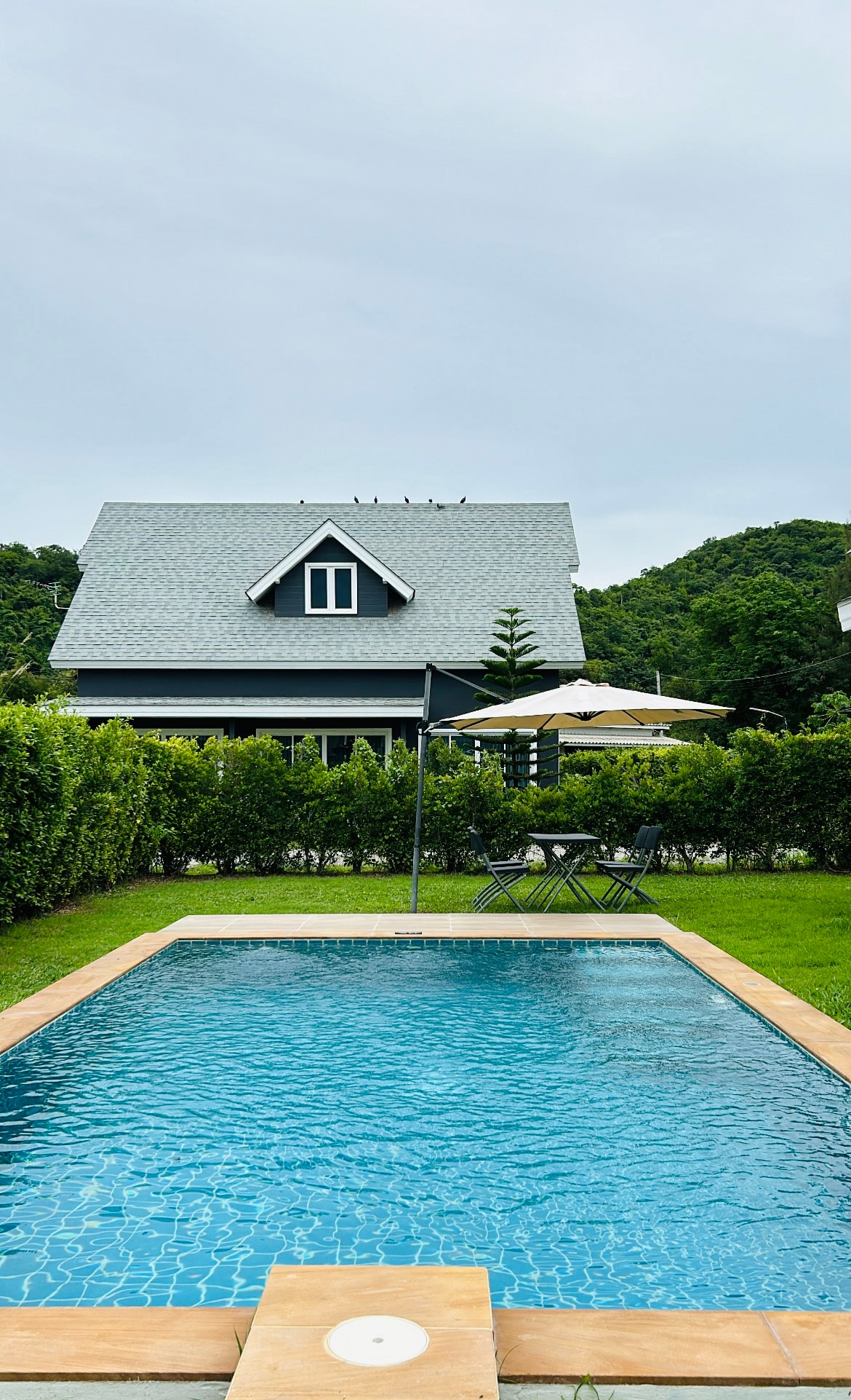 A private swimming pool is surrounded by well-maintained greenery, creating a serene environment. A stylish house with a gray roof is prominently positioned in the background. Outdoor seating and an umbrella are visible near the pool, enhancing the outdoor relaxation area.