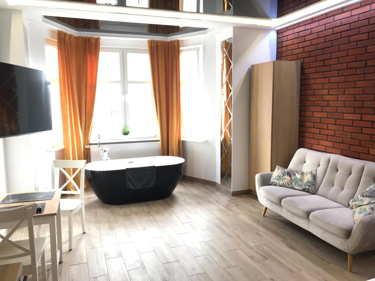 An open living space features a freestanding black bathtub as a focal point, surrounded by large windows adorned with orange curtains. A sleek television is mounted on the wall, while a light-colored sofa with decorative cushions complements a small table nearby.