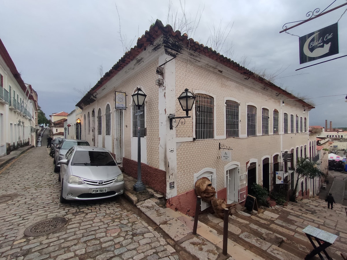 A corner building reflects colonial architecture with white walls adorned with decorative tiles. Street lamps are positioned along the cobblestone road, which gently slopes downward. A parked car is visible nearby, alongside details of surrounding greenery and a sign indicating a local establishment.