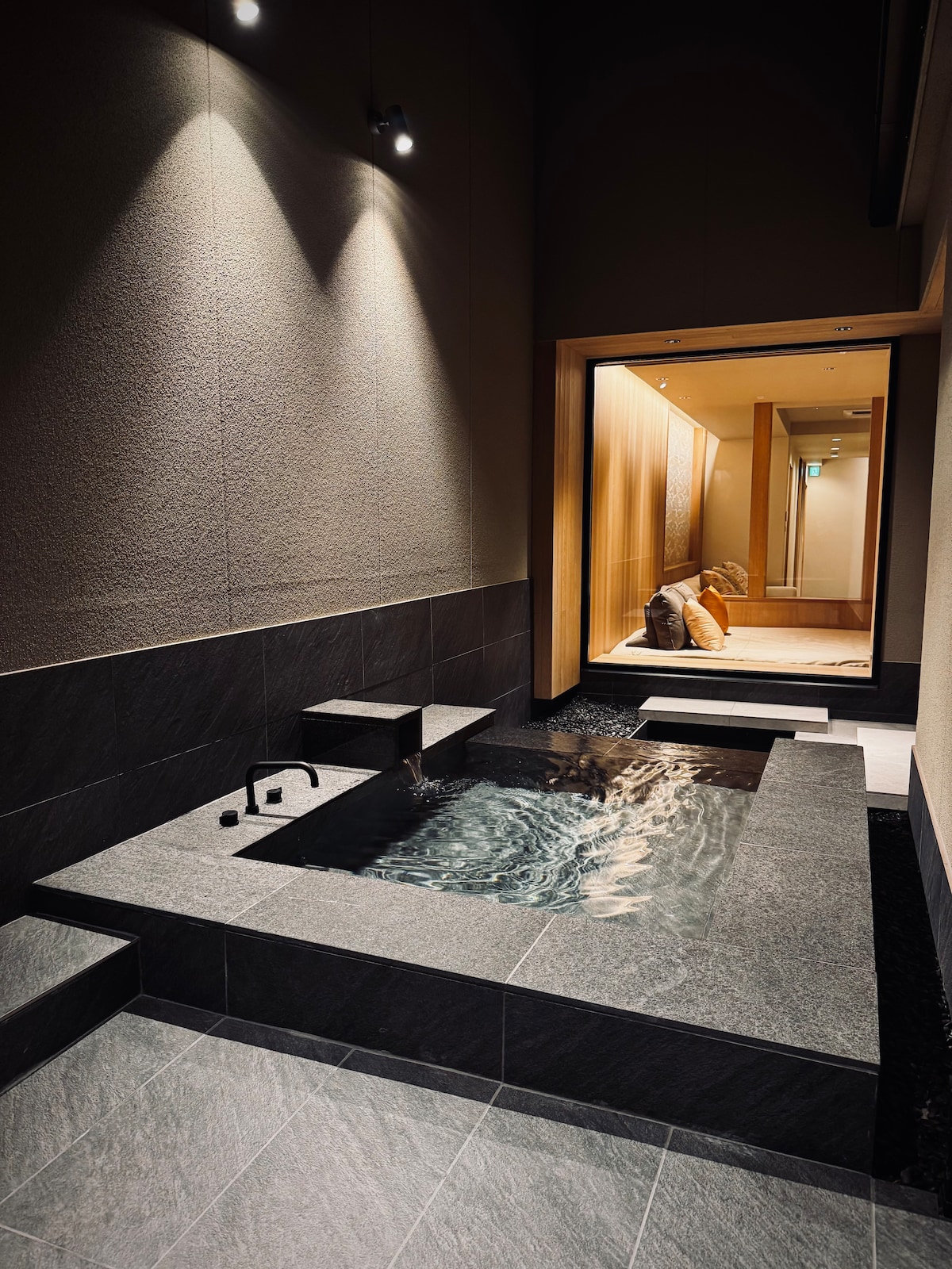 A modern outdoor bath featuring a sleek granite surface with a gentle water flow. Soft lighting highlights the elegant design, complemented by a corridor leading to a cozy seating area, enhancing the tranquil ambiance. Natural materials create a harmonious connection with the surroundings.