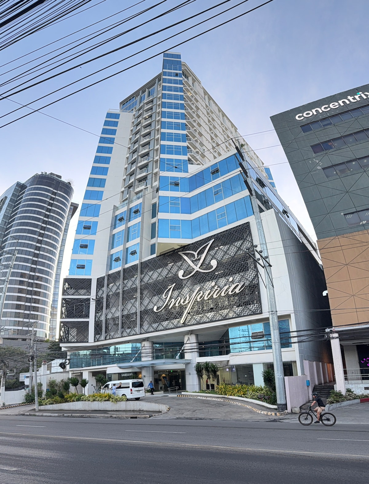A tall, modern building features a combination of blue and silver glass facades, with a prominent entrance showcasing an elegant sign. Surrounding landscaping includes manicured greenery, while nearby structures reflect contemporary architecture and city life.